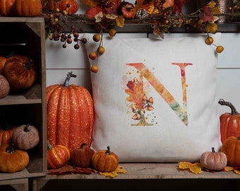 May include: A white throw pillow with a watercolor design of the letter N surrounded by fall leaves and acorns. The pillow is sitting on a wooden shelf with pumpkins and gourds.