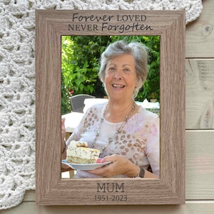 May include: A wooden photo frame with a light brown finish. The frame has a photo of a woman smiling and holding a piece of cake. The frame has the text "Forever Loved Never Forgotten" and "Mum 1951-2023" engraved on it.