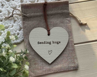 May include: A white wooden heart-shaped sign with the text "Sending hugs" and a small heart drawn below. The sign is hanging from a brown string and is displayed in a small burlap bag.