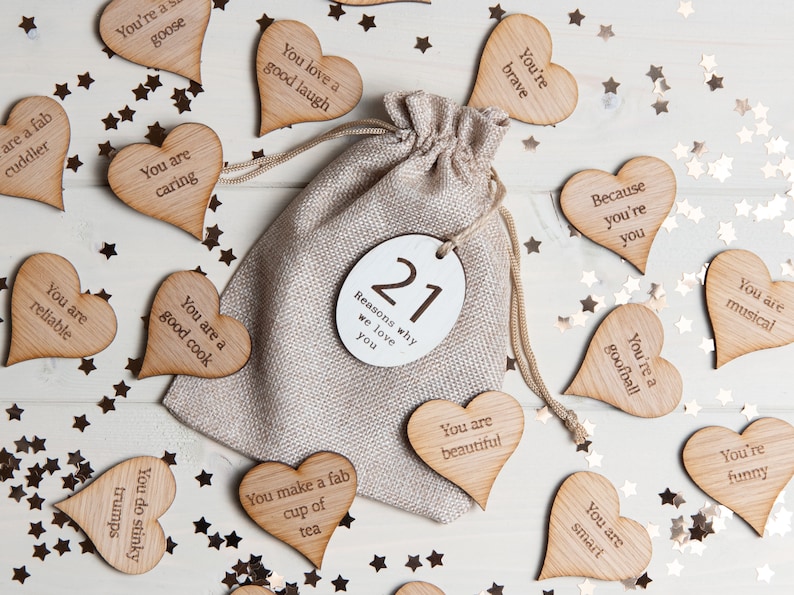 May include: A burlap bag filled with 21 wooden heart-shaped tokens, each with a reason why someone is loved. The bag is on a white wooden surface with gold glitter stars scattered around. The tokens have phrases like "You are a good cook" and "You are brave".