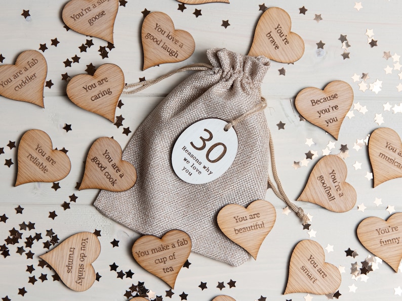 Puede incluir: Una bolsa de arpillera llena de 30 fichas de madera en forma de coraz&oacute;n, cada una con un mensaje positivo escrito en ella. La bolsa est&aacute; etiquetada con "30 razones por las que te queremos". Las fichas est&aacute;n esparcidas alrededor de la bolsa sobre una superficie de madera blanca con estrellas doradas brillantes.