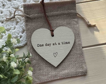 May include: A white wooden heart-shaped ornament with the text "One day at a time" and a small heart symbol. The ornament is hanging from a brown string and is in a burlap bag.