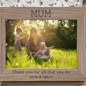 May include: A wooden photo frame with a light brown finish. The frame has a white mat and a clear glass front. The frame is decorated with the word "MUM" at the top and the text "Thank you for all that you do Love Ava & Sylvie xx" at the bottom. The frame contains a photo of a woman with two children in a grassy field.