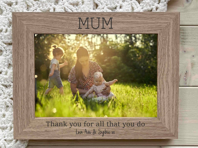 May include: A wooden photo frame with a light brown finish. The frame has a white mat and a clear glass front. The frame is decorated with the word "MUM" at the top and the text "Thank you for all that you do Love Ava & Sylvie xx" at the bottom. The frame contains a photo of a woman with two children in a grassy field.