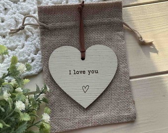 May include: A white wooden heart-shaped ornament with the text "I love you" and a small heart drawn below. The ornament is hanging from a brown string and is in a burlap bag.