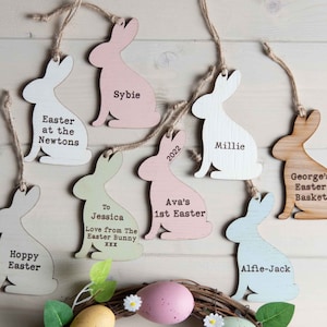 May include: Seven wooden bunny rabbit ornaments with personalised messages. The ornaments are painted in various pastel colours, including pink, green, blue, and white. The messages include names, ages, and Easter greetings.