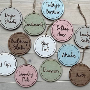 May include: A set of 12 round wooden tags with various labels, including "Dryer Sheets", "Condiments", "Bella's House", "Lonely Socks", "Building Blocks", "Your Text", "Vehicles", "Tips", "Laundry Pods", "Dinosaurs", "Herbs", and "Teddy's Birdbox".