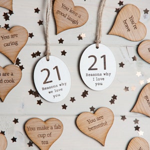 May include: Twenty-one wooden heart-shaped tags with various phrases written on them, including "You are caring", "You are a good cook", "You're brave", "You're a goofball", "You make a fab cup of tea", "You are beautiful", and "Because you're you". There are two white oval tags with the number "21" and the text "Reasons why we love you" and "Reasons why I love you". The tags are scattered on a white wooden surface with gold glitter stars.