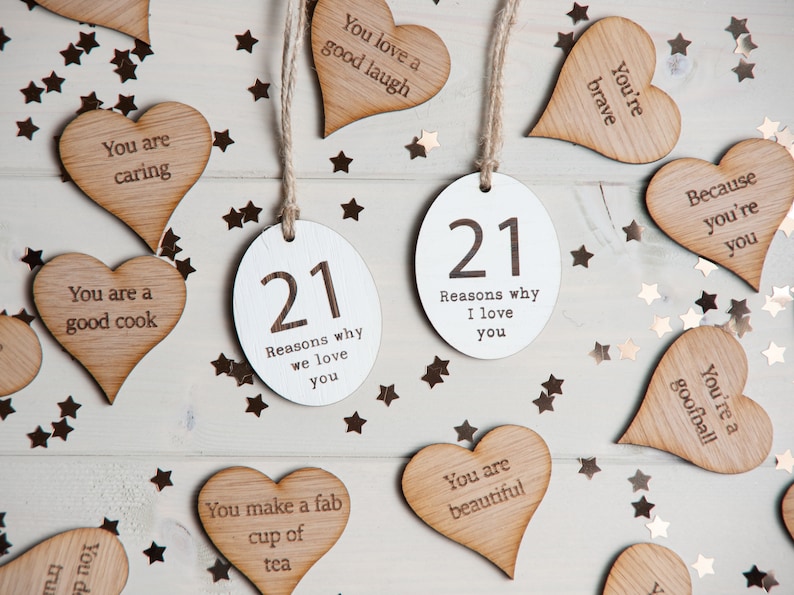 May include: Twenty-one wooden heart-shaped tags with various phrases written on them, including "You are caring", "You are a good cook", "You're brave", "You're a goofball", "You make a fab cup of tea", "You are beautiful", and "Because you're you". There are two white oval tags with the number "21" and the text "Reasons why we love you" and "Reasons why I love you". The tags are scattered on a white wooden surface with gold glitter stars.
