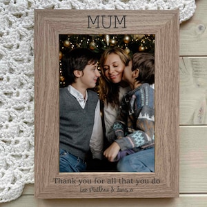 May include: A brown wooden photo frame with a picture of a mother and her two sons in front of a Christmas tree. The frame has the word "MUM" engraved at the top and the text "Thank you for all that you do Love Matthew & James xx" at the bottom.