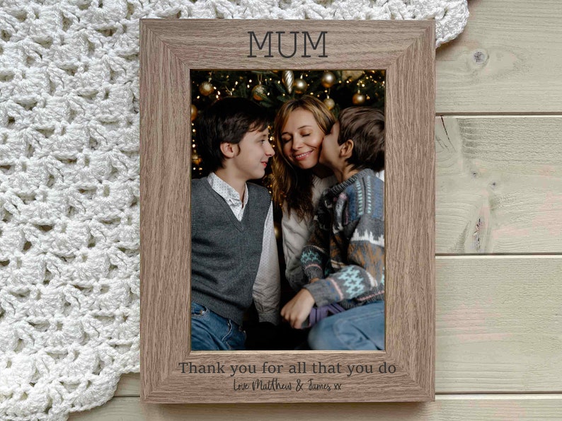 May include: A brown wooden photo frame with a picture of a mother and her two sons in front of a Christmas tree. The frame has the word "MUM" engraved at the top and the text "Thank you for all that you do Love Matthew & James xx" at the bottom.