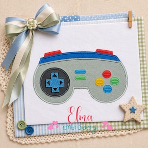 May include: A grey, blue, and red video game controller design. The controller features a directional pad, buttons, and a blue trim. The design is on a white background with a blue and green ribbon and a star-shaped embellishment. The text "Elma embroidery" is visible.