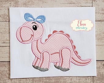 Könnte beinhalten: Rosa gestickter Dinosaurier mit einer blauen Schleife, stehend auf weißem Hintergrund. Der Dinosaurier hat ein Punkte-Muster und ein lächelndes Gesicht. Der Text "Elma embroidery" befindet sich in der oberen rechten Ecke des Bildes.