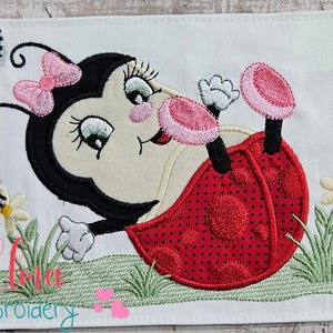 May include: Embroidered design of a red ladybug with black spots, pink wings, and black eyes. The ladybug is lying on its back with its legs in the air. The design is surrounded by green grass and a white daisy.