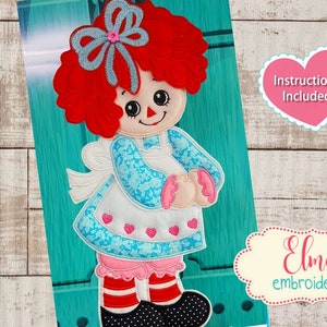 May include: An embroidered Raggedy Ann doll design with red yarn hair, a blue bow, and a blue floral dress. The design includes the text "Instructions Included" and is available in 5x7, 6x10, and 7x12 inch hoop sizes.