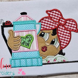 May include: Embroidered design of a cartoon woman with a red and white checkered bandana holding a blue and green coffee pot with a heart on the side. The design includes the text "Elma embroidery".