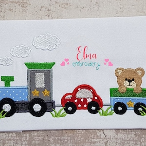 May include: Embroidered design of a toy train with a red car, a blue and green box car with a teddy bear peeking out, and white clouds. The text "Elma embroidery" is above the train.