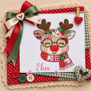 May include: A white fabric square featuring an embroidered patchwork reindeer wearing glasses and a scarf, with the name Elma embroidered below. The square is decorated with a red and green ribbon bow, a small wooden heart, and red buttons.