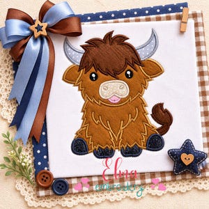 May include: A brown Highland cow applique with a smiling face, blue horns, and dark blue hooves. The design is on a white background, with the text "Elma embroidery" in pink script. Decorative ribbons and a star-shaped embellishment are also present.