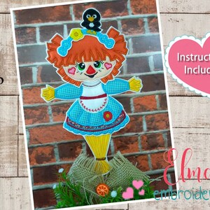May include: A red-haired scarecrow with a blue dress and a black bird on its head. The scarecrow is standing on a pile of straw. The image includes the text "Instructions Included" and "Elma Embroidery".