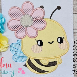Può includere: Disegno ricamato di un'ape di cartone animato carina con un fiore rosa sulla testa. L'ape ha strisce nere e gialle, ali blu e un viso sorridente. Il design è disponibile in varie dimensioni: 4x4, 5x4, 5x7, 5x8, 6x10 e 7x12 pollici.