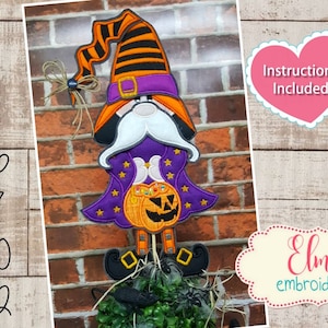 May include: Halloween gnome with a purple and orange hat, holding a pumpkin bucket with candy. The gnome is standing on a bed of green and black fabric. The image includes the text "ITH Hoop 5x7 6x10 7x12" and "Instructions Included" in a heart shape. The image also includes the text "Elma embroidery" in a cloud shape.