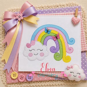 May include: A white fabric square featuring an embroidered rainbow with a smiling cloud, two butterflies, and the text Elma embroidery. The piece is decorated with a large lavender, yellow, and pink ribbon bow, a small heart clip, and a cloud applique.