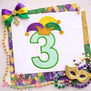 Peut inclure: Un fond blanc présente le chiffre 3 vert avec un motif en chevrons, surmonté d'un chapeau de bouffon et d'une couronne. Un nœud violet, or et vert avec un charme de fleur de lys est en haut à gauche. Des perles dorées et un masque sont également présents.