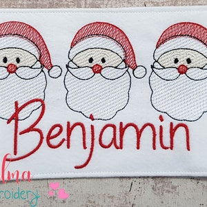 May include: White embroidered patch with three red-hatted Santa faces and the name "Benjamin" in red cursive lettering. The patch is surrounded by a white border.