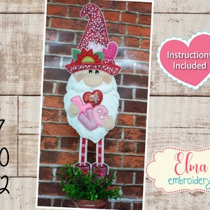 May include: A Valentine's Day gnome decoration with a red and white polka dot hat, holding a pink "love" sign. The gnome has a white beard and is decorated with hearts and flowers. The sign says "Instructions Included" and "Elma embroidery".