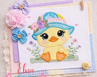 May include: A charming embroidered duckling wearing a blue hat adorned with flowers, sitting amidst embroidered greenery and flowers. The duckling is yellow with orange feet and a matching beak. The image includes decorative bows and the text "Elma embroidery".