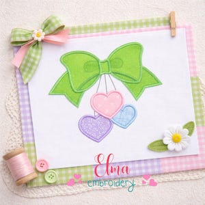 Puede incluir: Un dise&ntilde;o de bordado decorativo con un gran lazo verde y tres corazones colgantes en rosa, azul y morado. El dise&ntilde;o est&aacute; sobre un fondo blanco con un borde de cuadros vichy en verde, rosa y morado. Tambi&eacute;n est&aacute; presente el texto "Elma embroidery".