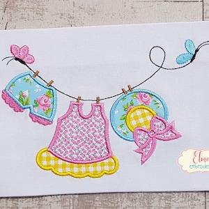 Puede incluir: Diseño bordado de un tendedero con pantalones cortos de estampado floral azul y blanco, un vestido de estampado floral rosa y amarillo y un sombrero de estampado floral azul y blanco con un lazo a cuadros amarillo y blanco. El diseño incluye dos mariposas.