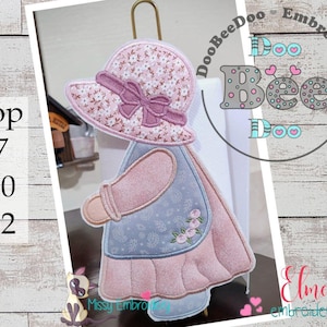 May include: An embroidered girl doll with a pink hat and a floral design. The doll is holding a paper towel roll. The image includes the text "DooBeeDoo Embroidery" and "Elma Embroidery".