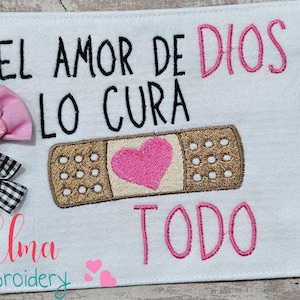 May include: White fabric with pink and brown embroidery. The text "EL AMOR DE DIOS LO CURA TODO" is embroidered in pink and brown. The text "Elma embroidery" is embroidered in pink at the bottom. The sizes 4x4, 5x4, 5x7, 5x8, 6x10, and 7x12 are listed on the right side of the fabric.