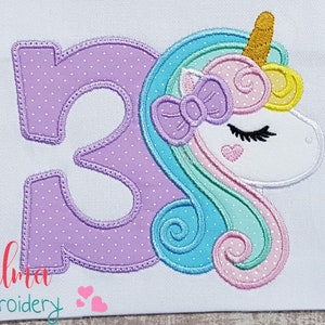 May include: A white fabric applique featuring a unicorn design with the number 3. The number 3 is lavender with white polka dots. The unicorn has a gold horn, a lavender bow, and a pink heart. The text "Elma embroidery" is at the bottom.