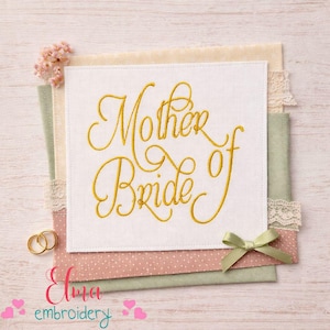 May include: A white square embroidered with the words "Mother of Bride" in gold script. The embroidery is on a stack of fabric in shades of cream, green, and pink, with lace trim and a green ribbon. Two gold rings and a small flower arrangement are also present.