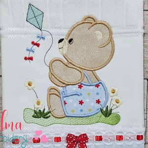 May include: Embroidered design of a brown bear wearing blue overalls with white stars and red accents, holding a blue kite with red ribbons. The bear is sitting on green grass with white flowers. The design is on a white fabric with a white lace trim and red bows.