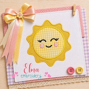 May include: A cheerful, embroidered sun design with a yellow gingham pattern, smiling face, and pink cheeks. The design is on a white background, accented with a pink and purple patterned paper, a decorative bow, and the text "Elma embroidery".