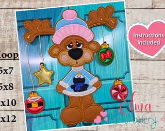 May include: Embroidered design of a reindeer wearing a blue and pink hat and sweater, holding a penguin. The reindeer has antlers with four hanging ornaments: a gold star, a red ornament with a Santa hat, a green ornament with a red bow, and a red and white candy cane ornament. The text "Instructions Included" is in a pink heart shape.