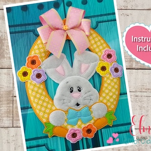 Può includere: Una ghirlanda pasquale con un volto di coniglietto, carote e fiori colorati. La ghirlanda ha un motivo floreale giallo e un fiocco rosa. Il testo "Instructions Included" è in un cuore rosa.