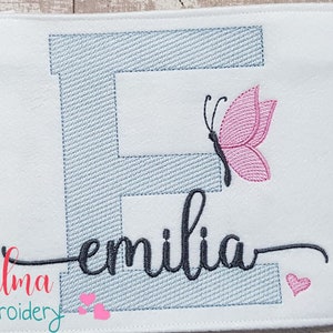 May include: Embroidered design featuring a blue letter E with a pink butterfly and the name "emilia" in black cursive script. The design is on a white fabric background.