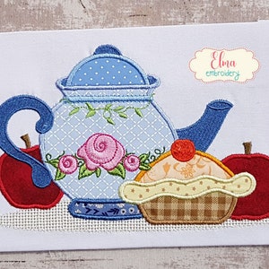 May include: Embroidered design of a blue teapot with a floral pattern, two red apples, and a yellow cupcake with a red cherry on top. The design is on a white fabric background. The text "Elma embroidery" is in the top right corner.
