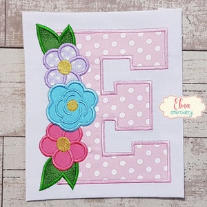 May include: Embroidered letter E with pink polka dot background and a cluster of flowers in shades of purple, blue, and pink with green leaves.