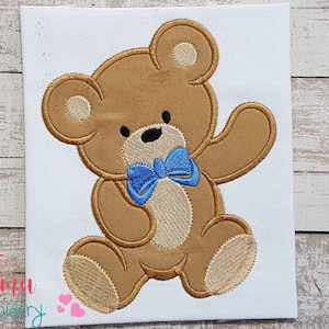 May include: Embroidered design of a brown teddy bear wearing a blue bow tie. The bear is standing with its right arm raised.