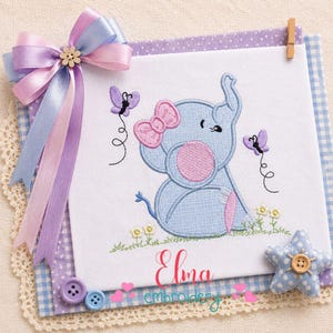 May include: A white fabric square featuring an embroidered light blue elephant with a pink bow and ear, surrounded by two purple butterflies and small flowers. The piece is decorated with a lavender and blue ribbon bow, buttons, and the text Elma embroidery.