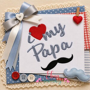 May include: A white fabric square featuring embroidered text that reads I heart my Papa in blue, with a red heart icon and a black mustache graphic. The square is decorated with a blue and white satin bow, a small red heart clip, and three buttons.