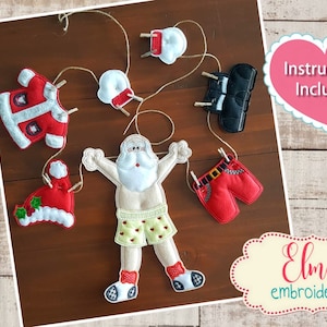 Pode incluir: Um conjunto de enfeites de Natal de feltro para um projeto de bordado no aro. Os enfeites incluem um Papai Noel, um chapéu, uma jaqueta, uma calça e um par de meias. Os enfeites estão pendurados em uma corda com prendedores de roupa. O texto "Instruções Incluídas" está em um coração rosa. O texto "Elma bordado" está em um oval creme com borda vermelha.
