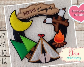 May include: A black embroidery hoop with a wreath design featuring a yellow crescent moon, green trees, a brown tent with footprints, a brown stick with white marshmallows, and an orange fire. The wreath is decorated with a brown banner that says "Happy Camper".