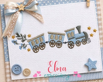 May include: A white card with a blue train embroidery design, accented with gold stars and floral elements. The card is decorated with a blue and tan bow, buttons, and a star-shaped embellishment. The name "Elma" is written in pink cursive.